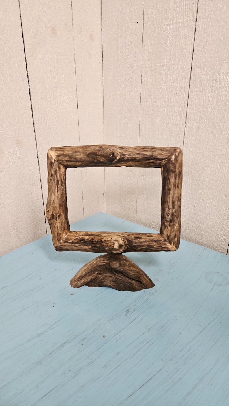 May include: A rustic, rectangular wooden picture frame with a natural, unfinished appearance. The frame is supported by a triangular base, and the wood has visible knots and grain. The frame sits on a light blue surface.