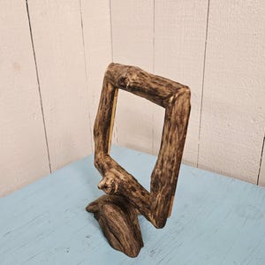 May include: A rustic, wooden picture frame made from a natural branch. The frame is a square shape with a rough, textured surface and a dark brown color. It sits on a light blue surface against a white wooden background.