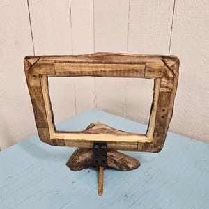 May include: A rustic wooden picture frame with a natural, unfinished look. The frame is rectangular with a wide border, supported by a wooden base and a metal bracket. The frame is displayed on a light blue surface.