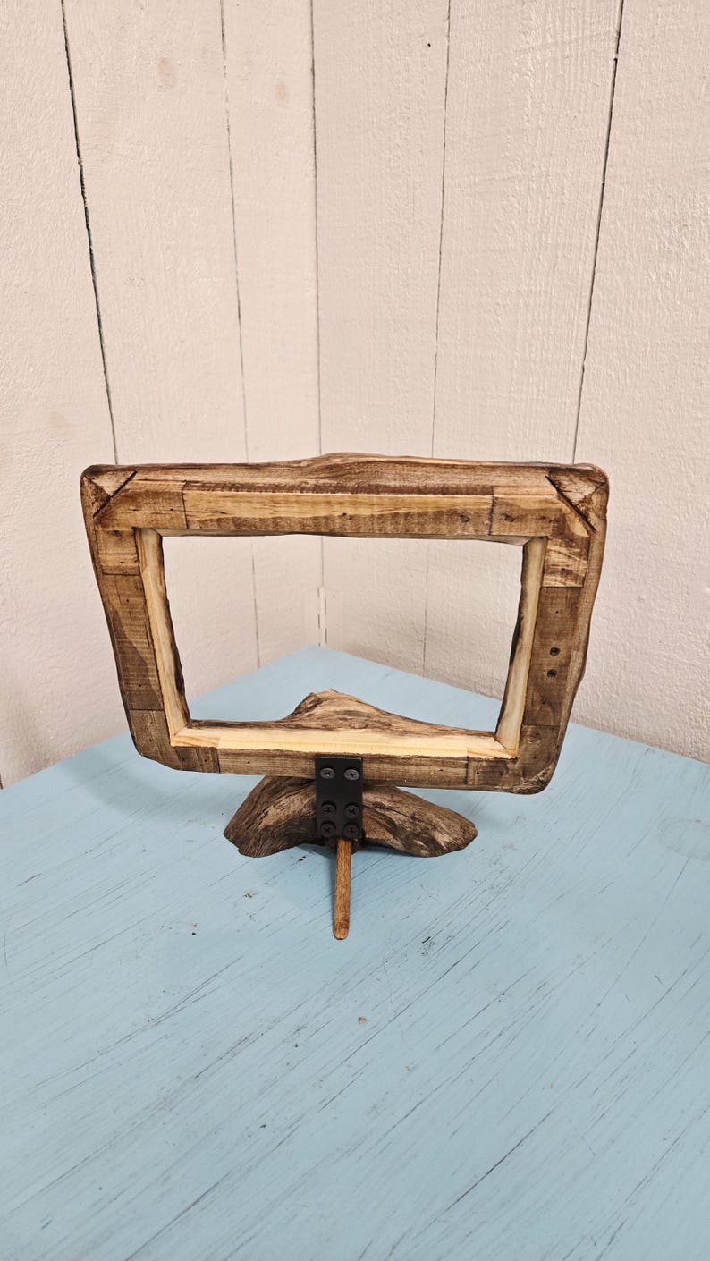 May include: A rustic wooden picture frame with a natural, unfinished look. The frame is rectangular with a wide border, supported by a wooden base and a metal bracket. The frame is displayed on a light blue surface.