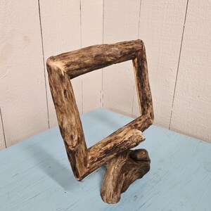 May include: A rustic, square wooden frame made from natural driftwood. The frame is angled, with a base that appears to be a continuation of the wood. The wood has a natural, weathered appearance, with varying shades of brown and tan.
