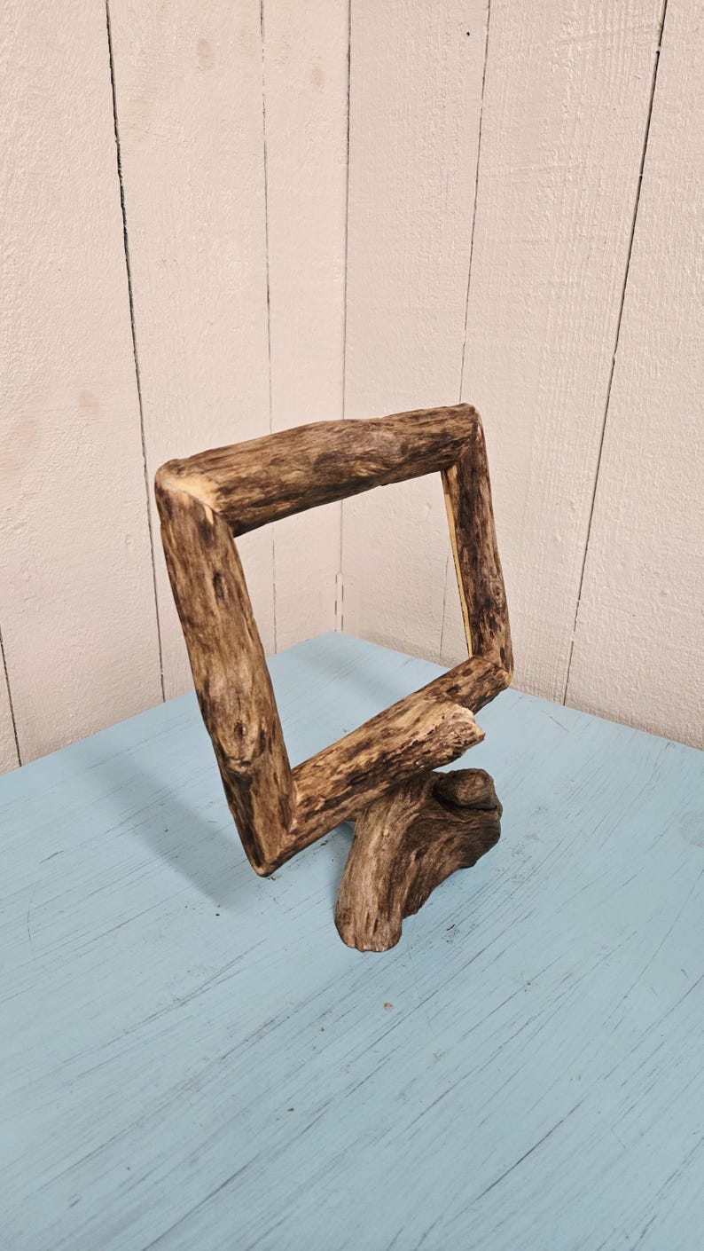 May include: A rustic, square wooden frame made from natural driftwood. The frame is angled, with a base that appears to be a continuation of the wood. The wood has a natural, weathered appearance, with varying shades of brown and tan.