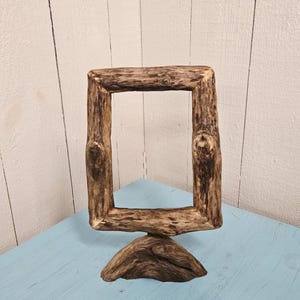 May include: A rustic, square wooden picture frame made from natural branches. The frame is brown with visible knots and a rough texture, standing on a wooden base. The frame is displayed on a light blue surface.
