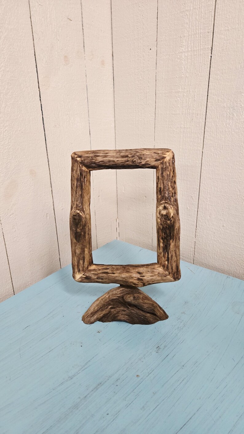 May include: A rustic, square wooden picture frame made from natural branches. The frame is brown with visible knots and a rough texture, standing on a wooden base. The frame is displayed on a light blue surface.