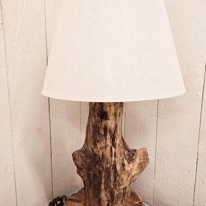 Peut inclure: Une lampe de table rustique en bois flotté avec un abat-jour en tissu blanc. La lampe est sur une base en bois et a un cordon.
