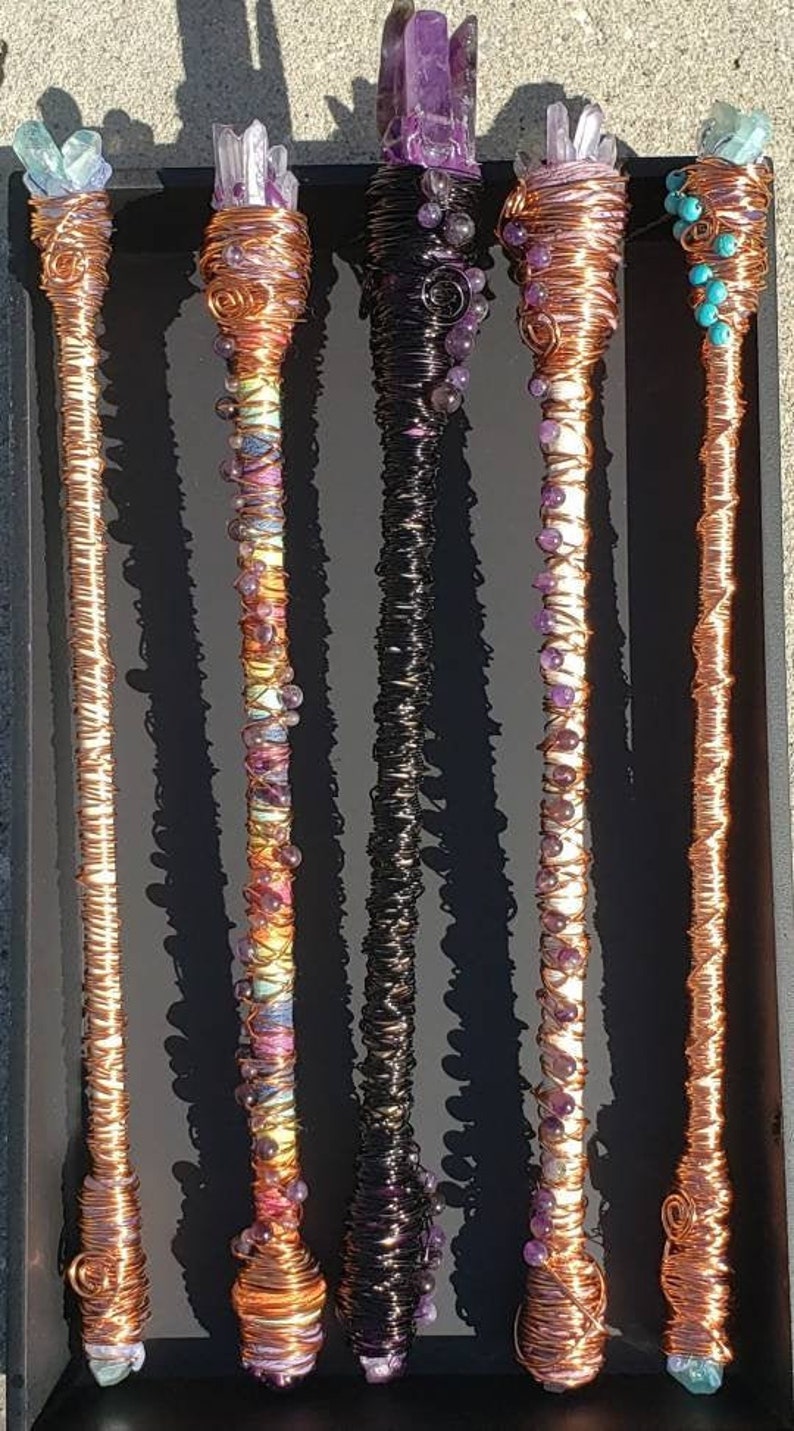 May include: Five copper wire wrapped wands with various colored crystals and beads. The wands are displayed on a black surface.