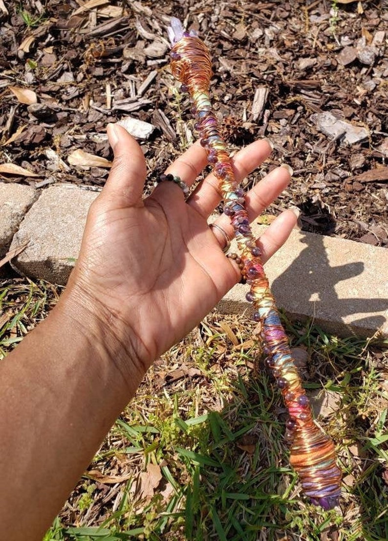 May include: A copper wire wrapped wand with amethyst crystals and colorful beads. The wand is held in a hand.