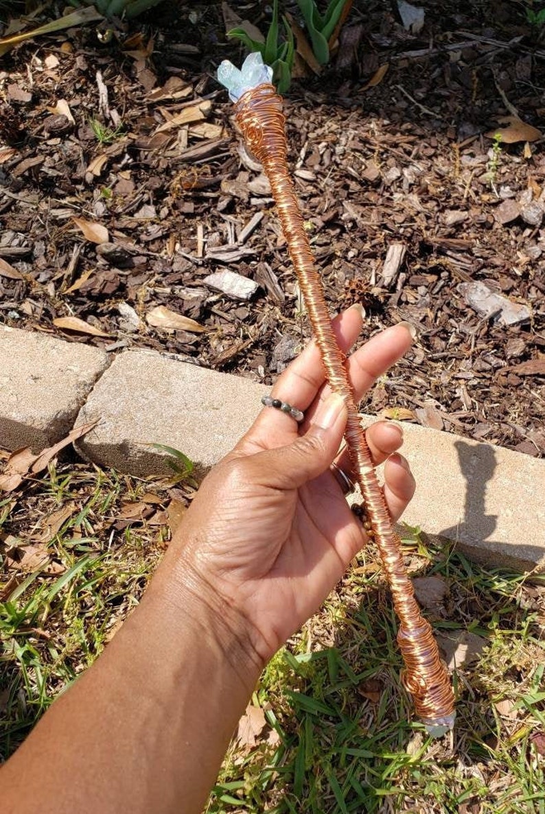May include: A copper wire wrapped wand with a light blue crystal at the top. The wand is held in a hand.