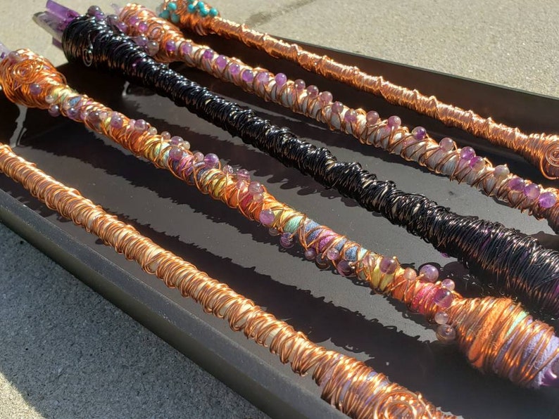 May include: Five copper wire wrapped wands with amethyst and other colorful beads. The wands are displayed on a black surface.