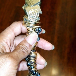 May include: A gold and black beaded wand with a large pyrite crystal at the top. The wand is wrapped in wire and has black beads interspersed throughout.