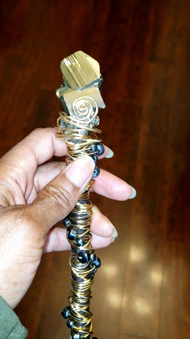 May include: A gold and black beaded wand with a large pyrite crystal at the top. The wand is wrapped in wire and has black beads interspersed throughout.