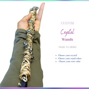 May include: A custom crystal wand wrapped in gold wire with black beads. The wand is held in a hand. The text "CUSTOM Crystal Wands (MADE TO ORDER)" is visible. Choose your crystal, wand colors, and wire color.