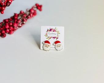 Puede incluir: Un par de pendientes de botón de plata con un rostro de Papá Noel de dibujos animados con un gorro rojo, barba blanca y ojos negros. Los pendientes están en una tarjeta blanca con el texto "Creations by Cathy" impreso en ella.