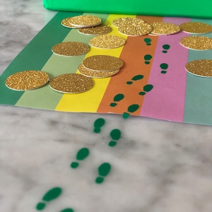 May include: A rainbow-colored path with green footprints leading away from a box filled with gold glitter circles.