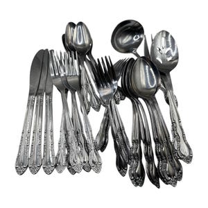 Puede incluir: Un conjunto de cubiertos de acero inoxidable, que incluye cuchillos, tenedores, cucharas, un cucharón y una cuchara ranurada. La cubertería presenta un motivo floral decorativo en los mangos. Los utensilios están dispuestos sobre un fondo blanco.