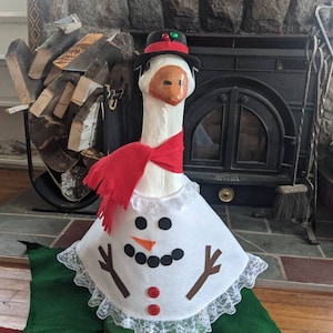 May include: A white goose dressed as a snowman with a red scarf, black top hat with a green pom-pom, and a white snowman costume with brown twig arms and red buttons.