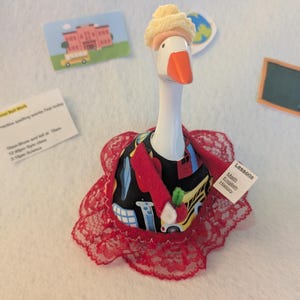 May include: A whimsical goose figurine dressed as a school teacher. The goose wears a black dress with school bus and building graphics, a red jacket, and a red lace collar. A small paper reads "Lessons: Math, English, History."