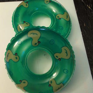 May include: Two inflatable green pool floats with yellow duck designs. The floats are round with a hole in the center. The ducks are facing different directions around the float. The floats are on a white surface.