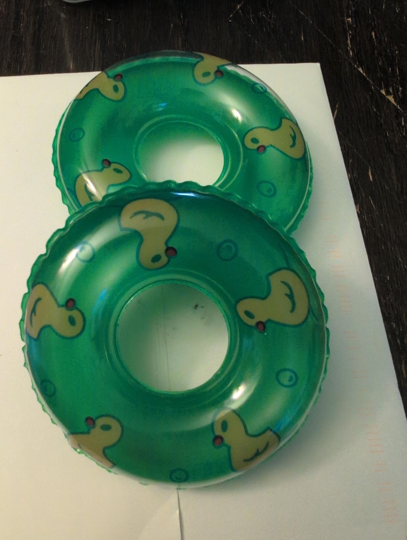 May include: Two inflatable green pool floats with yellow duck designs. The floats are round with a hole in the center. The ducks are facing different directions around the float. The floats are on a white surface.