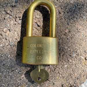 May include: A brass padlock with a U-shaped shackle and a key inserted into the bottom. The front of the lock body is engraved with the text COLONIAL PIPELINE CO. The metal surface has a brushed finish and shows minor signs of wear and use.