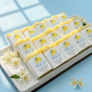 May include: A tray of lemon-scented soaps, each wrapped with a label featuring a lemon illustration and the words "Thank You." The soaps are arranged on a white tray, with white flowers and a light blue background.