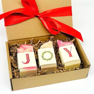 May include: A gift box with three bars of soap arranged inside, with the letters "JOY" printed on the front. The soaps are wrapped in paper with decorative designs and are nestled in brown paper. A red ribbon is tied on top.