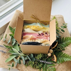 May include: A brown cardboard box tied with gold string, filled with six colorful bars of soap. The soaps are various colors, including pink, white, and blue, and are decorated with dried flowers and other natural elements. The box is surrounded by greenery.