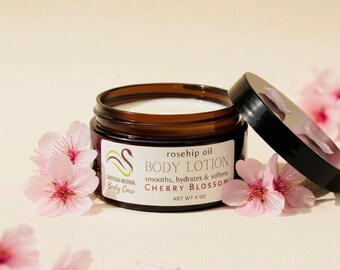 May include: A jar of cherry blossom body lotion with the lid off, surrounded by pink cherry blossoms. The label reads "rosehip oil BODY LOTION" and "NET WT 4 OZ". The jar is brown glass.