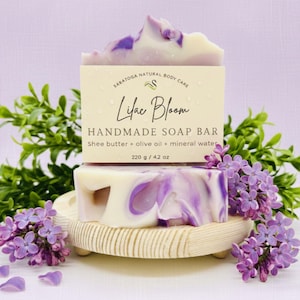 May include: A handmade soap bar labeled "Lilac Bloom" with a cream and purple swirl design. The bar is made with shea butter, olive oil, and mineral water. The soap is 4.2 oz (119 g) and is displayed with lilac flowers and greenery.