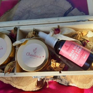 Puede incluir: Una caja de madera con tapa abatible contiene dos peque&ntilde;os frascos de crema facial y una botella de suero facial. Los frascos y la botella est&aacute;n etiquetados con "Chanita's Naturals". La botella de suero est&aacute; etiquetada con "Rose Silk Anti-Aging Face Serum" y tiene una etiqueta rosa con texto blanco. La caja est&aacute; llena de flores secas y otros materiales naturales.