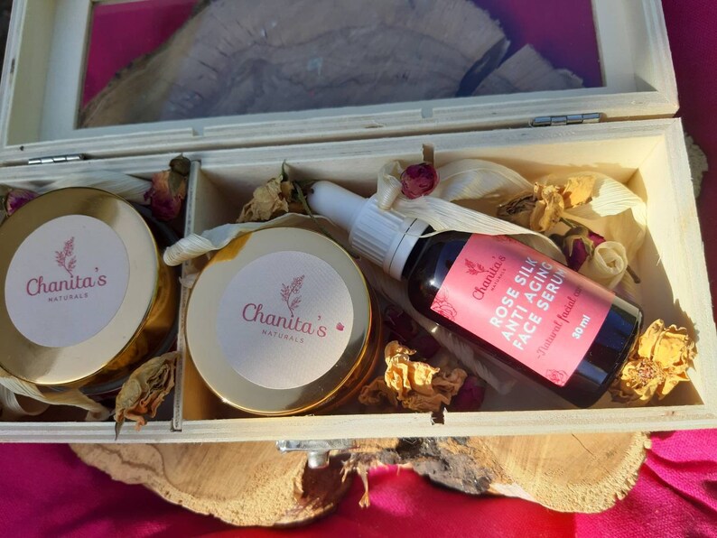 Puede incluir: Una caja de madera con tapa abatible contiene dos peque&ntilde;os frascos de crema facial y una botella de suero facial. Los frascos y la botella est&aacute;n etiquetados con "Chanita's Naturals". La botella de suero est&aacute; etiquetada con "Rose Silk Anti-Aging Face Serum" y tiene una etiqueta rosa con texto blanco. La caja est&aacute; llena de flores secas y otros materiales naturales.