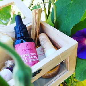 Puede incluir: Una caja de madera llena de productos de belleza naturales, que incluyen una botella de suero facial antienvejecimiento de seda de rosa, una pastilla de jab&oacute;n, un b&aacute;lsamo labial y un palo de incienso. La caja est&aacute; abierta y se encuentra sobre un fondo verde frondoso con una flor morada.