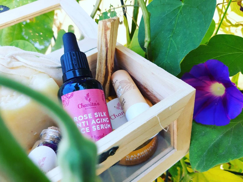 Puede incluir: Una caja de madera llena de productos de belleza naturales, que incluyen una botella de suero facial antienvejecimiento de seda de rosa, una pastilla de jab&oacute;n, un b&aacute;lsamo labial y un palo de incienso. La caja est&aacute; abierta y se encuentra sobre un fondo verde frondoso con una flor morada.