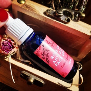 Puede incluir: Un peque&ntilde;o frasco de vidrio azul con un gotero blanco, que contiene un suero facial con aroma a rosa. La etiqueta dice "Suero facial antiedad de rosa - Hecho a mano natural - 30 ml".