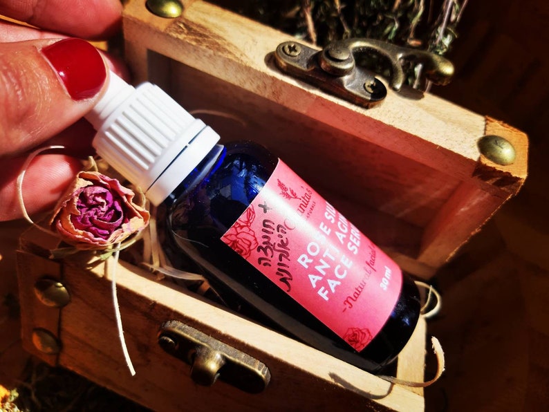 Puede incluir: Un peque&ntilde;o frasco de vidrio azul con un gotero blanco, que contiene un suero facial con aroma a rosa. La etiqueta dice "Suero facial antiedad de rosa - Hecho a mano natural - 30 ml".