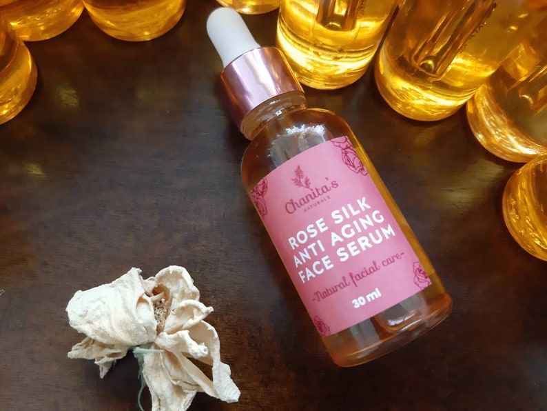 Puede incluir: Una botella de vidrio de suero facial antienvejecimiento de seda de rosa con una etiqueta rosa y un gotero. La etiqueta dice "Chanita's Naturals Rose Silk Anti-Aging Face Serum - Natural facial care - 30 ml".