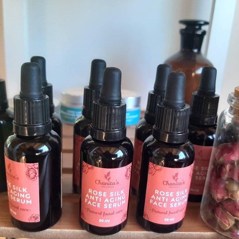 May include: Six small glass bottles with black dropper tops filled with a clear liquid. The bottles have labels with the text "Chanita's Naturals Rose Silk Anti-Aging Face Serum - Natural Facial Care - 30 ml".