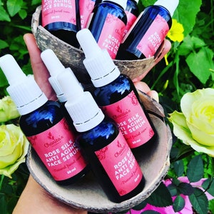 Puede incluir: Seis peque&ntilde;os frascos de vidrio azul con goteros blancos y etiquetas que dicen "Chanita's Rose Silk Anti Aging Face Serum - Natural facial care - 30 ml" est&aacute;n dispuestos en una c&aacute;scara de coco. Los frascos est&aacute;n en una mano, y el fondo es una imagen borrosa de rosas rosas y blancas.