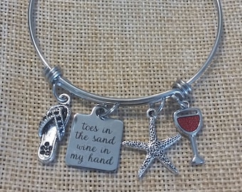 May include: A silver bangle bracelet with charms. The charms include a flip-flop, a wine glass, a starfish, and a square charm with the text "toes in the sand wine in my hand".