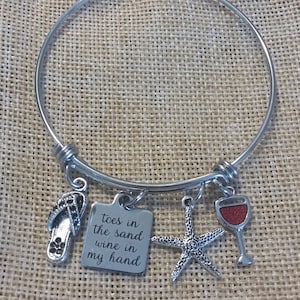 May include: A silver bangle bracelet with charms. The charms include a flip-flop, a wine glass, a starfish, and a square charm with the text "toes in the sand wine in my hand".
