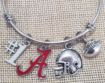 May include: A silver bangle bracelet with charms. The charms include a silver football, a silver helmet, a silver number one, and a red letter A.