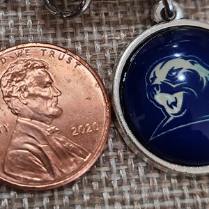 May include: A silver-colored pendant with a blue background and a white panther logo next to a 2020 penny. The pendant is round and has a shiny, domed surface. The penny is copper-colored and shows the profile of Abraham Lincoln.