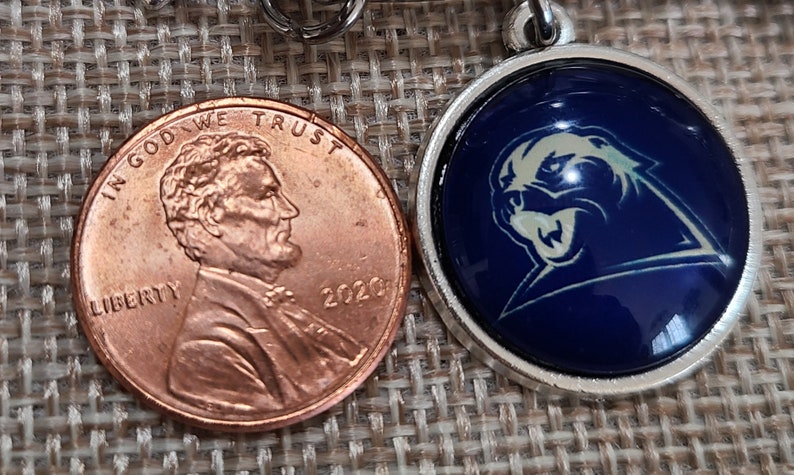 May include: A silver-colored pendant with a blue background and a white panther logo next to a 2020 penny. The pendant is round and has a shiny, domed surface. The penny is copper-colored and shows the profile of Abraham Lincoln.