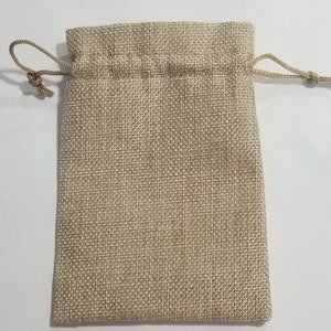 May include: A beige burlap drawstring bag. The bag is rectangular with a woven texture. The top of the bag has a drawstring closure made of a matching beige cord. The bag is empty and sits on a white surface.