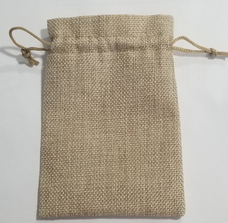 May include: A beige burlap drawstring bag. The bag is rectangular with a woven texture. The top of the bag has a drawstring closure made of a matching beige cord. The bag is empty and sits on a white surface.