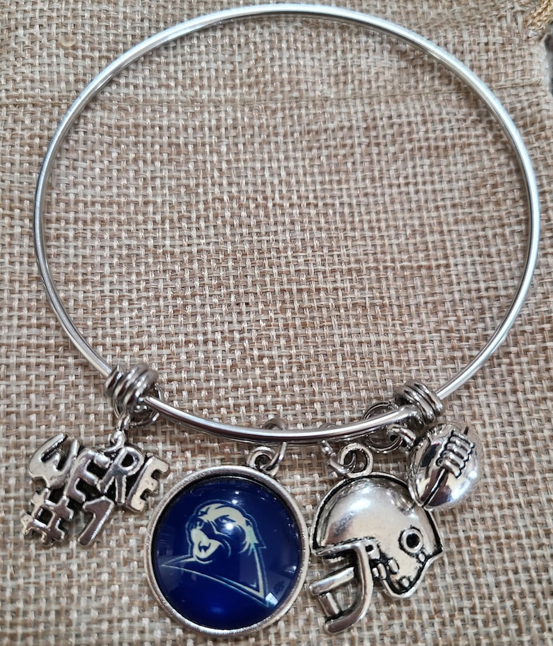 May include: Silver-toned bangle bracelet with multiple charms. Charms include a football helmet, a football, a round charm with a blue background and a panther logo, and a charm that says "WE'RE #1".