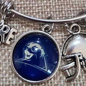 May include: Silver-toned charm bracelet with football-themed charms. Charms include a football helmet, a football, and a round charm with a blue and white logo. Additional charms spell out "WE'RE #7".