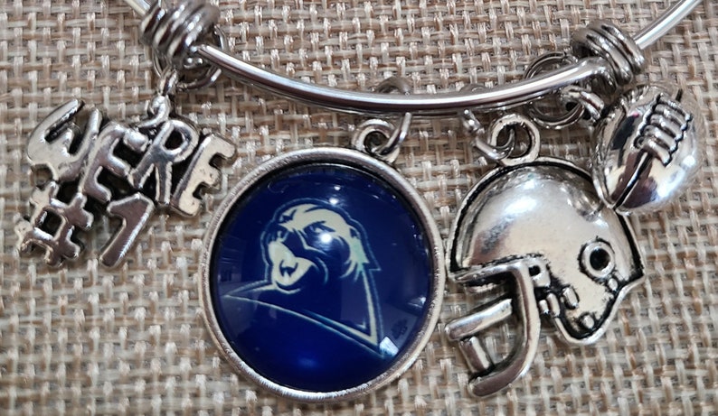 May include: Silver-toned charm bracelet with football-themed charms. Charms include a football helmet, a football, and a round charm with a blue and white logo. Additional charms spell out "WE'RE #7".