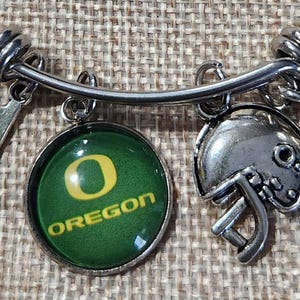 May include: A silver-toned charm bracelet featuring multiple charms. Charms include a football, a football helmet, a green and yellow Oregon Ducks logo, and a number one charm. The bracelet is on a textured, tan surface.