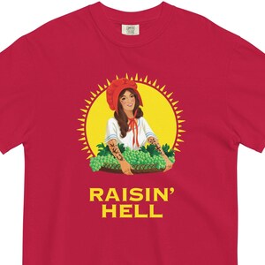 Puede incluir: Camiseta roja con un gráfico de una mujer con un gorro rojo sosteniendo uvas, sobre un fondo de rayos de sol amarillos. El texto "RAISIN' HELL" está impreso en amarillo debajo.